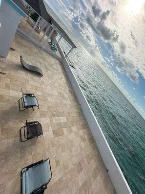 Een zonnig buitengebied met betegelde vloer, loungestoelen en uitzicht op de oceaan.