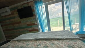 Een kamer met een bed, een aan de muur bevestigde televisie en uitzicht op de zee door een raam met blauwe gordijnen.