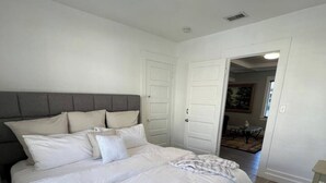 Ein Schlafzimmer mit einem großen Bett, weißer Bettwäsche und einem grauen Kopfteil. Zwei weiße Türen und ein kleiner Tisch mit einer Lampe sind vorhanden.