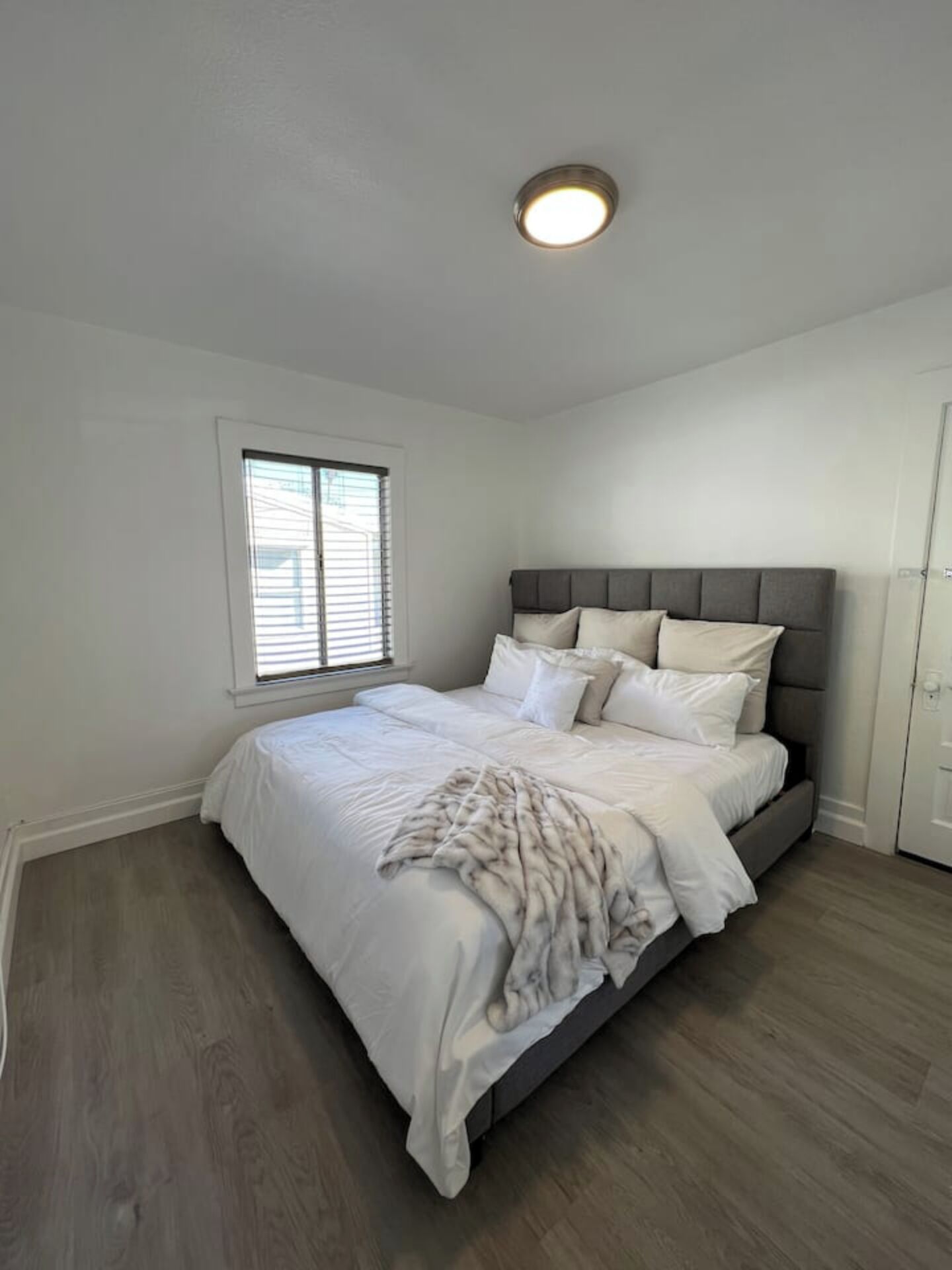 A bedroom with a bed, a window with blinds, a ceiling light, and a door.