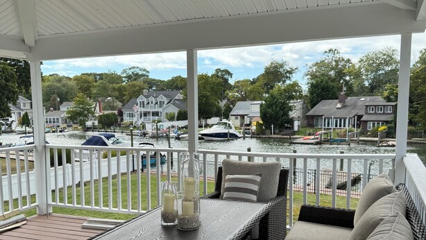 Een overdekt terras met rieten meubilair en een tafel, met uitzicht op een jachthaven met boten en huizen.