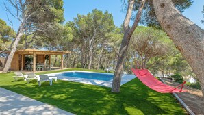 A backyard with a swimming pool, green lawn, and a wooden pergola with chairs.