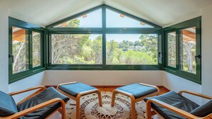 A room with a high ceiling and a large bay window overlooking a lush landscape. There are three wooden chairs with cushions and a woven rug on the floor.
