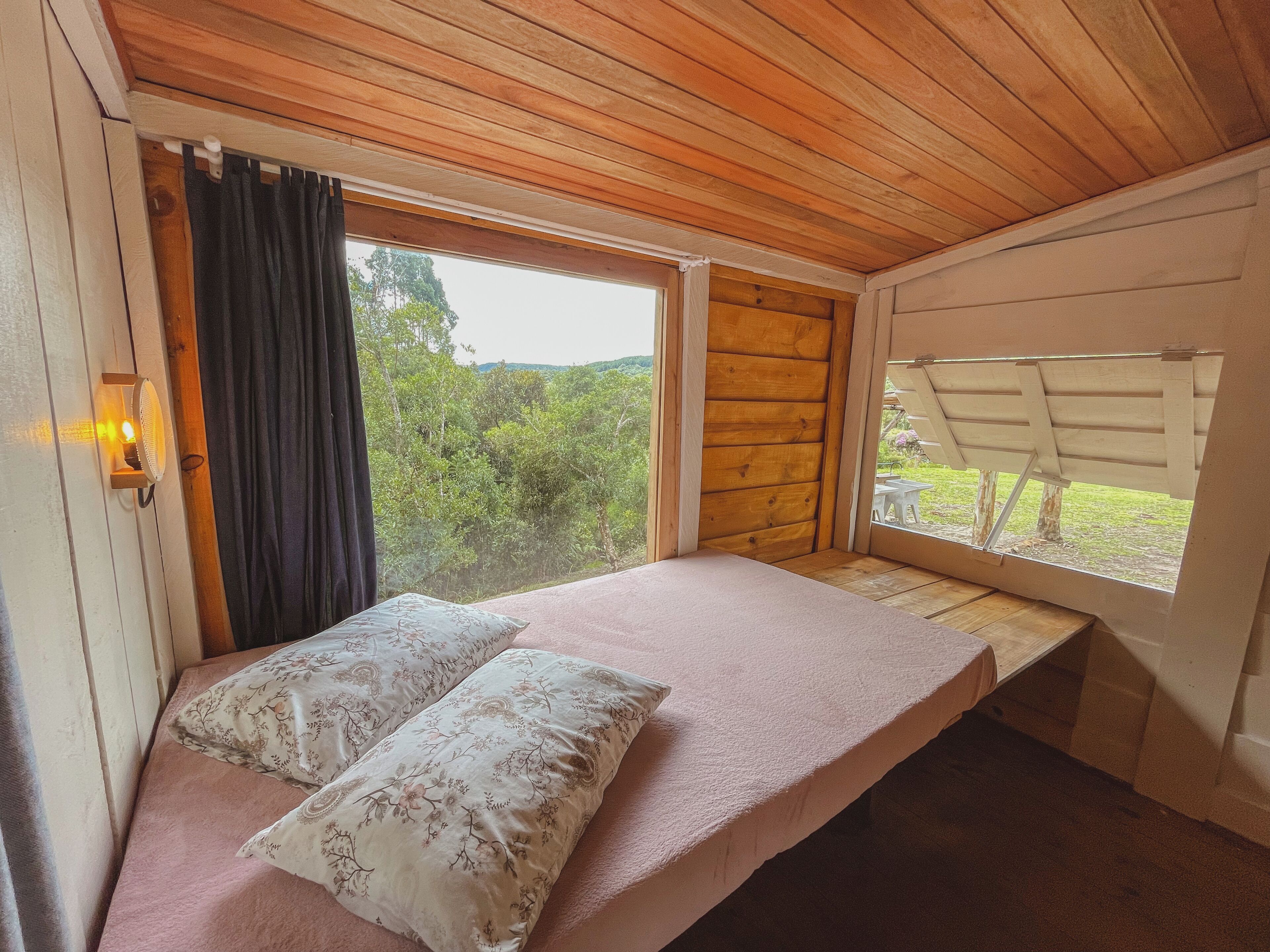 Un dormitorio con techo de madera, una cama con almohadas florales, una ventana con vistas al verdor y una mesita junto a la ventana.