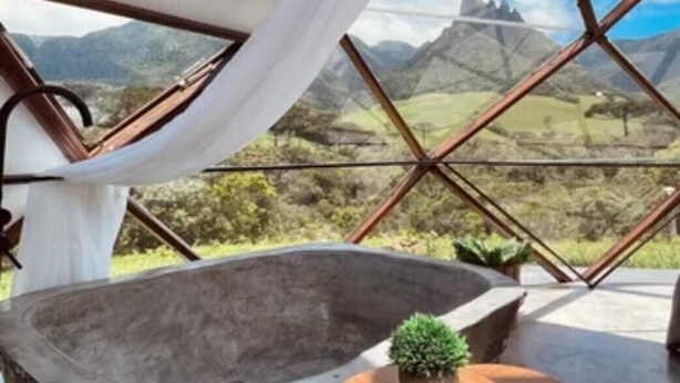 Une pièce dotée d’une grande baignoire en pierre, d’une petite table avec une plante et offrant une vue sur les montagnes à travers une structure de fenêtre aux motifs géométriques.