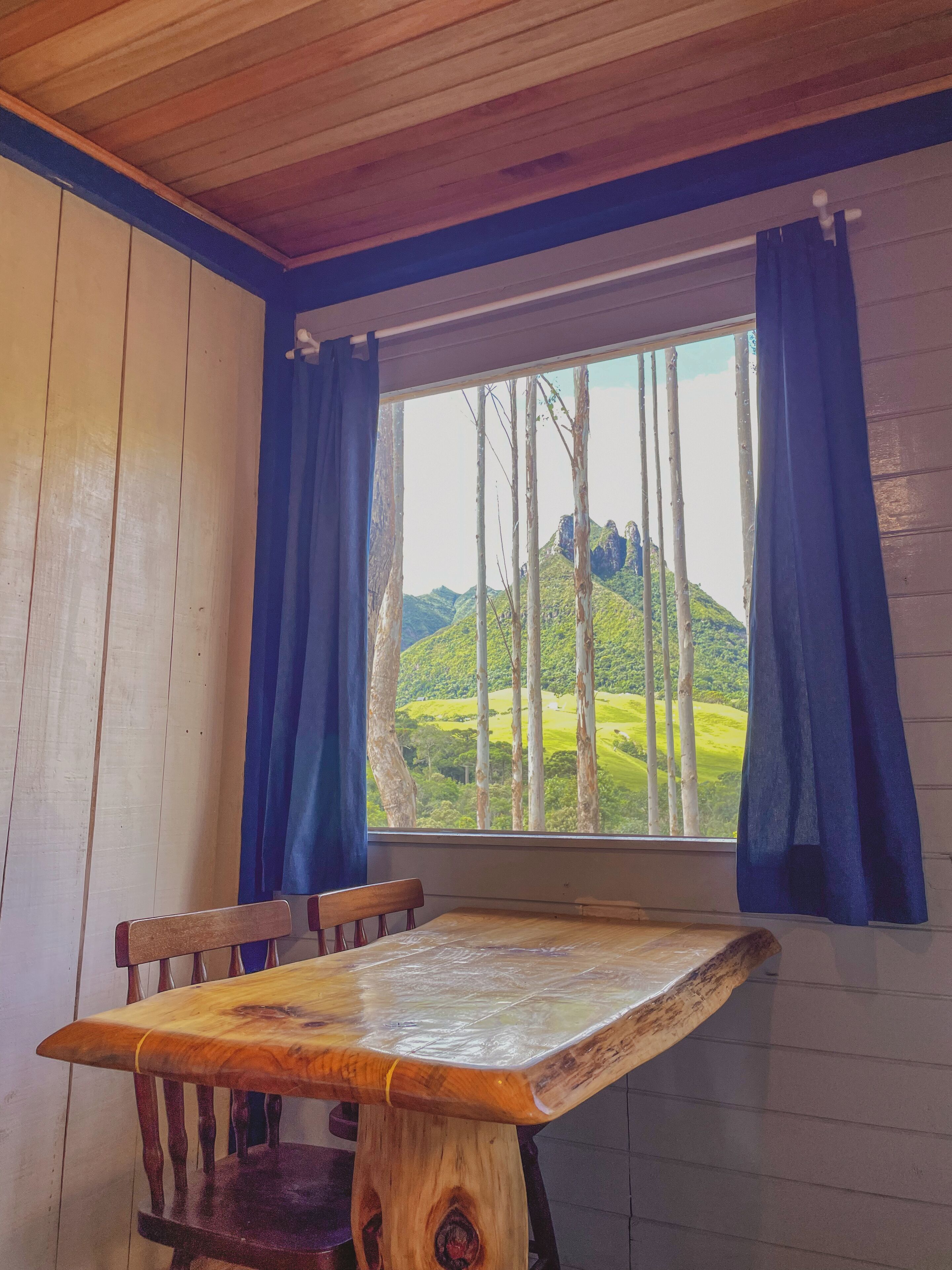 Una mesa de madera con sillas, una ventana con cortinas azules y vistas a colinas y árboles verdes.