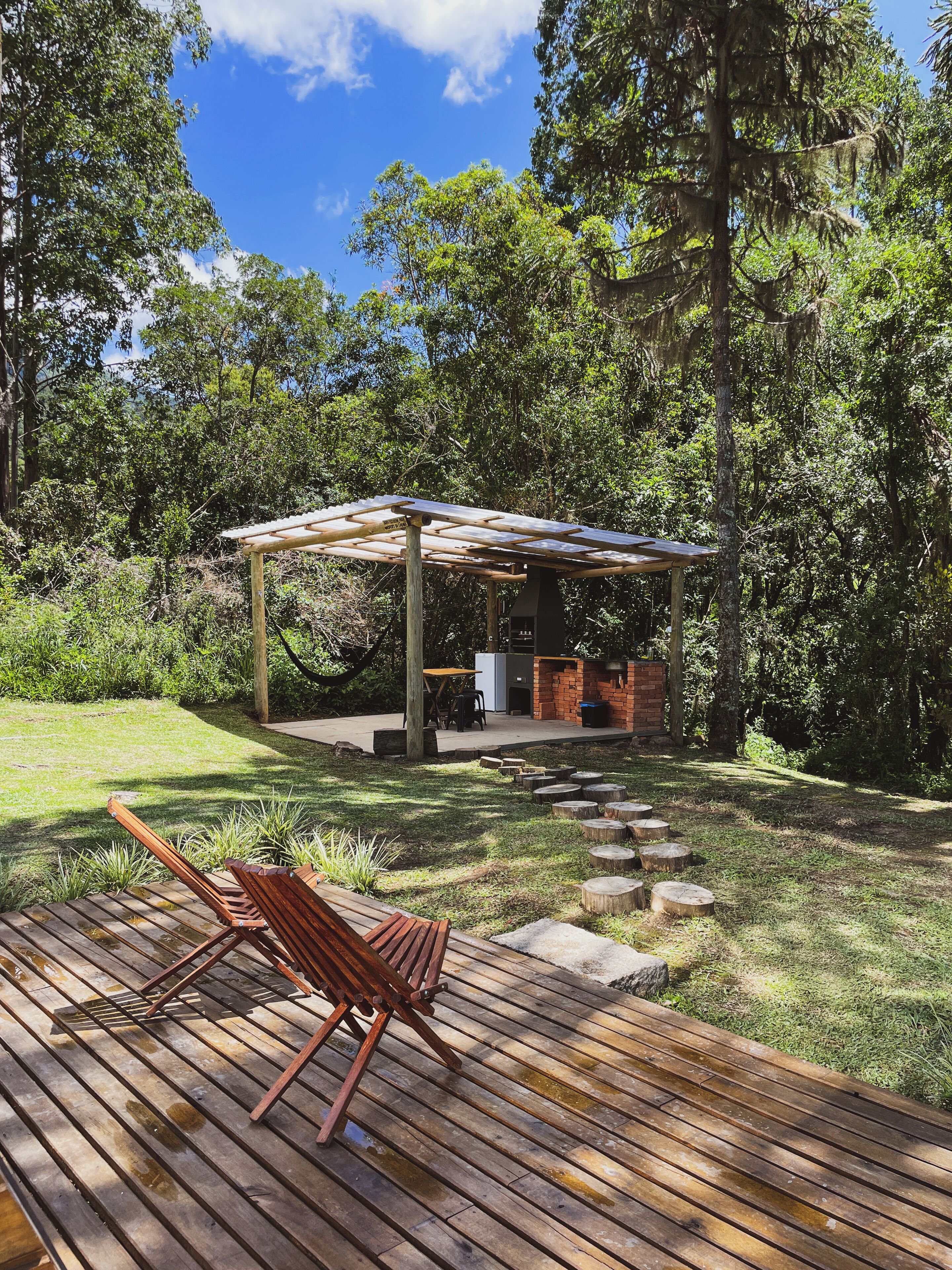 Une terrasse en bois agrémentée d’une pergola, d’un espace barbecue en briques et d’un coin salon avec un banc en bois.