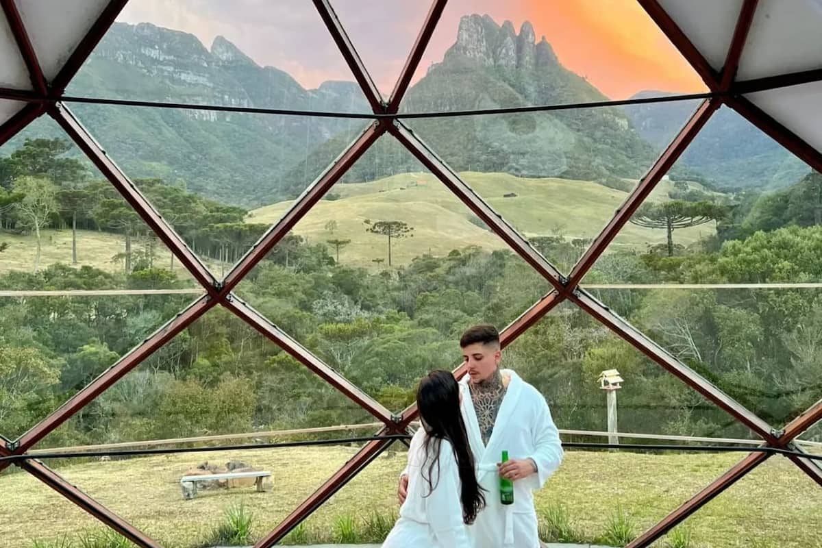 A couple sitting on a bed inside a geodesic dome structure with a view of a mountainous landscape.