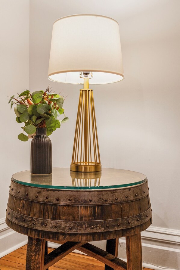 Una mesa redonda de madera con superficie de vidrio, una lámpara moderna con pantalla blanca y un jarrón con follaje verde.