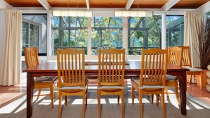 A dining area with a wooden table and chairs, large windows with a view of trees, and a ceiling with wooden beams. - Home Sweet Holiday in Lilli Pilli (Lilli Pilli)
