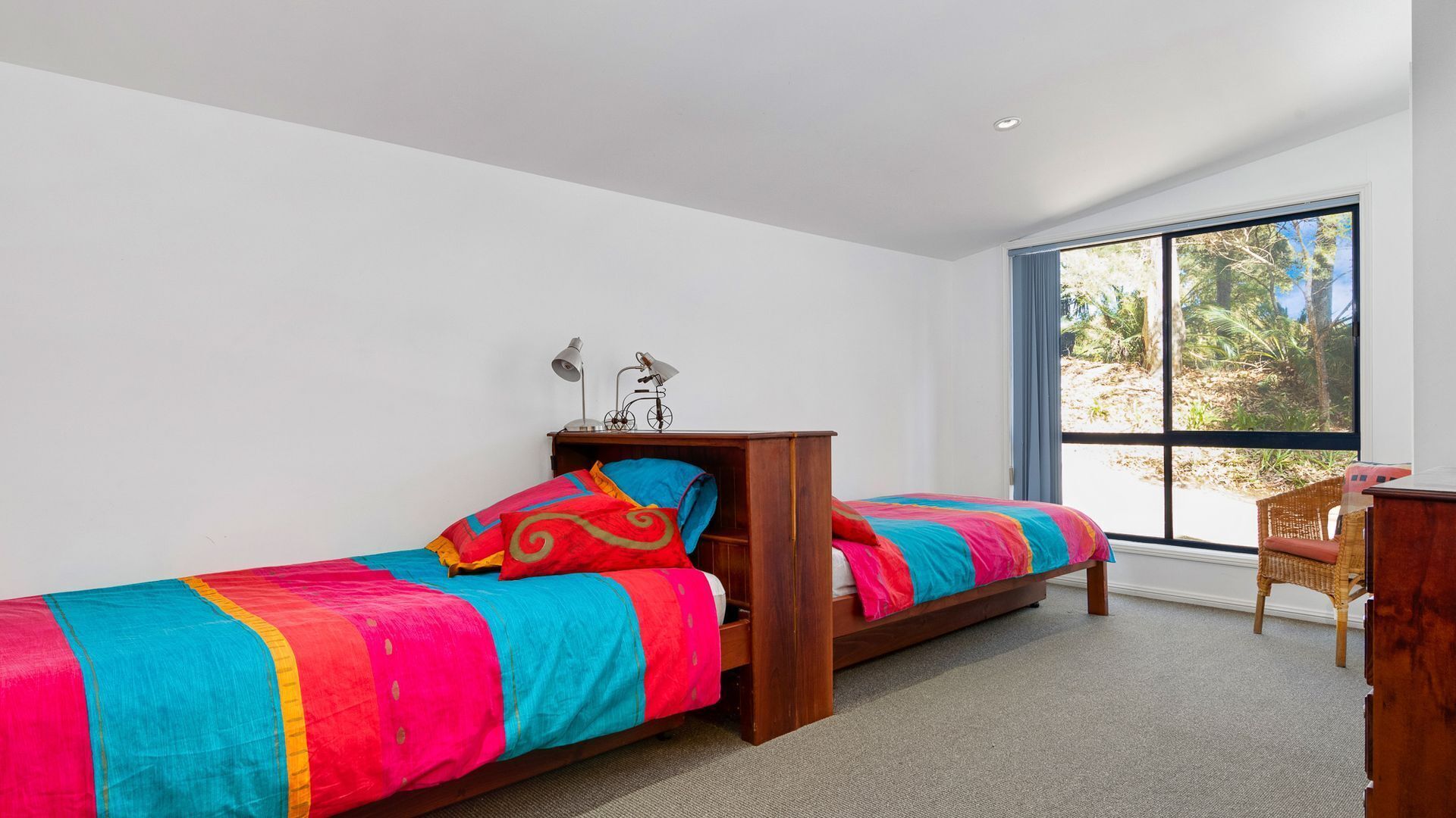 Un dormitorio con dos camas individuales, una mesita de noche con lámparas, una silla de mimbre y un ventanal con vistas a zonas verdes.