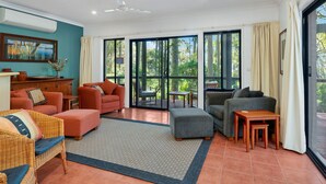 A spacious living room with a large sliding glass door leading to an outdoor deck, furnished with sofas, a coffee table, and a wicker chair. - Home Sweet Holiday in Lilli Pilli (Lilli Pilli)