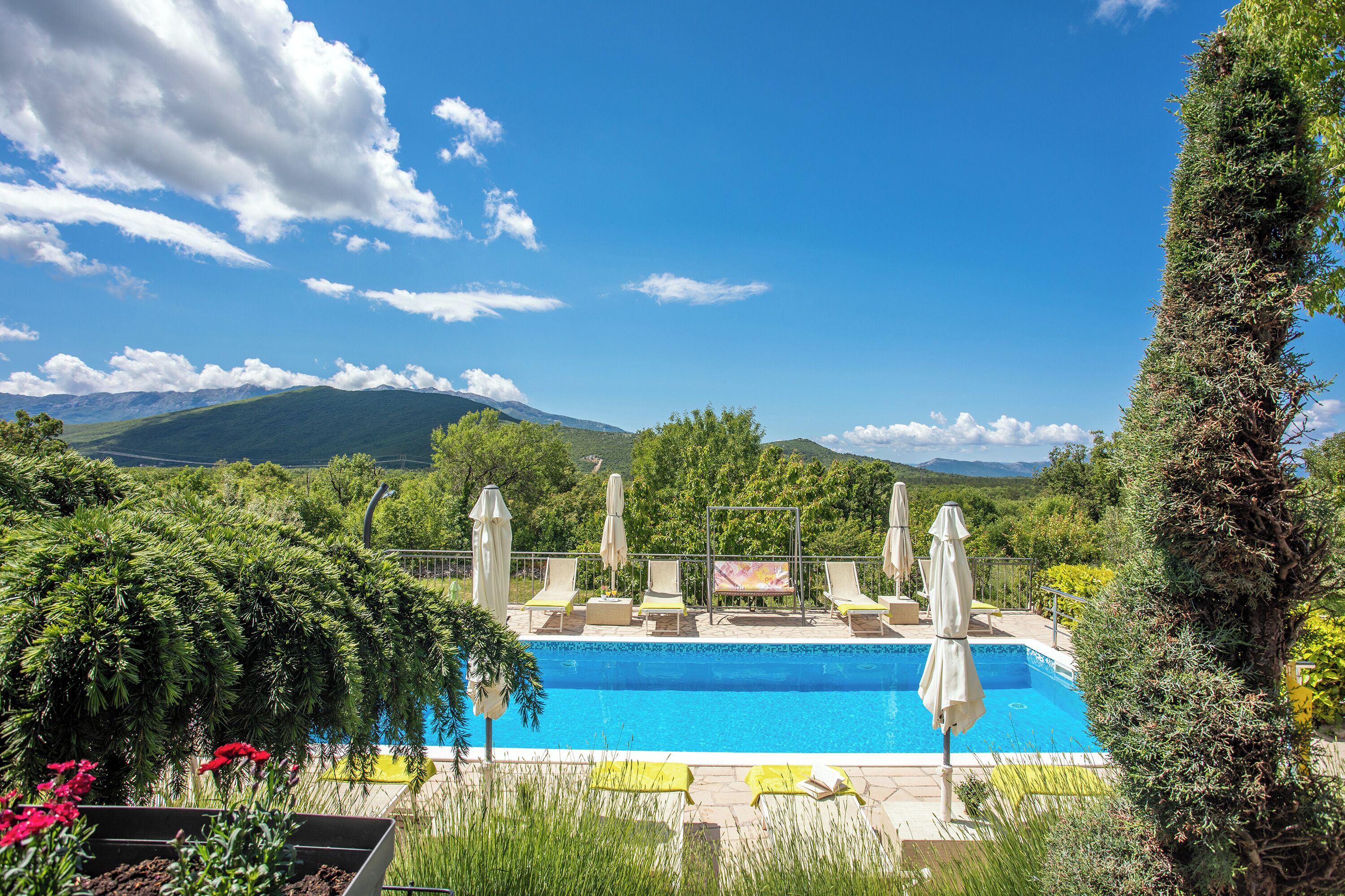 A swimming pool with lounge chairs and umbrellas, surrounded by greenery and mountains in the background.