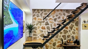 Eine Treppe mit einer Steinwand, ein Esstisch mit Stühlen, ein Blumentopf und ein an der Wand befestigtes Fernsehgerät, das eine Strandansicht zeigt.