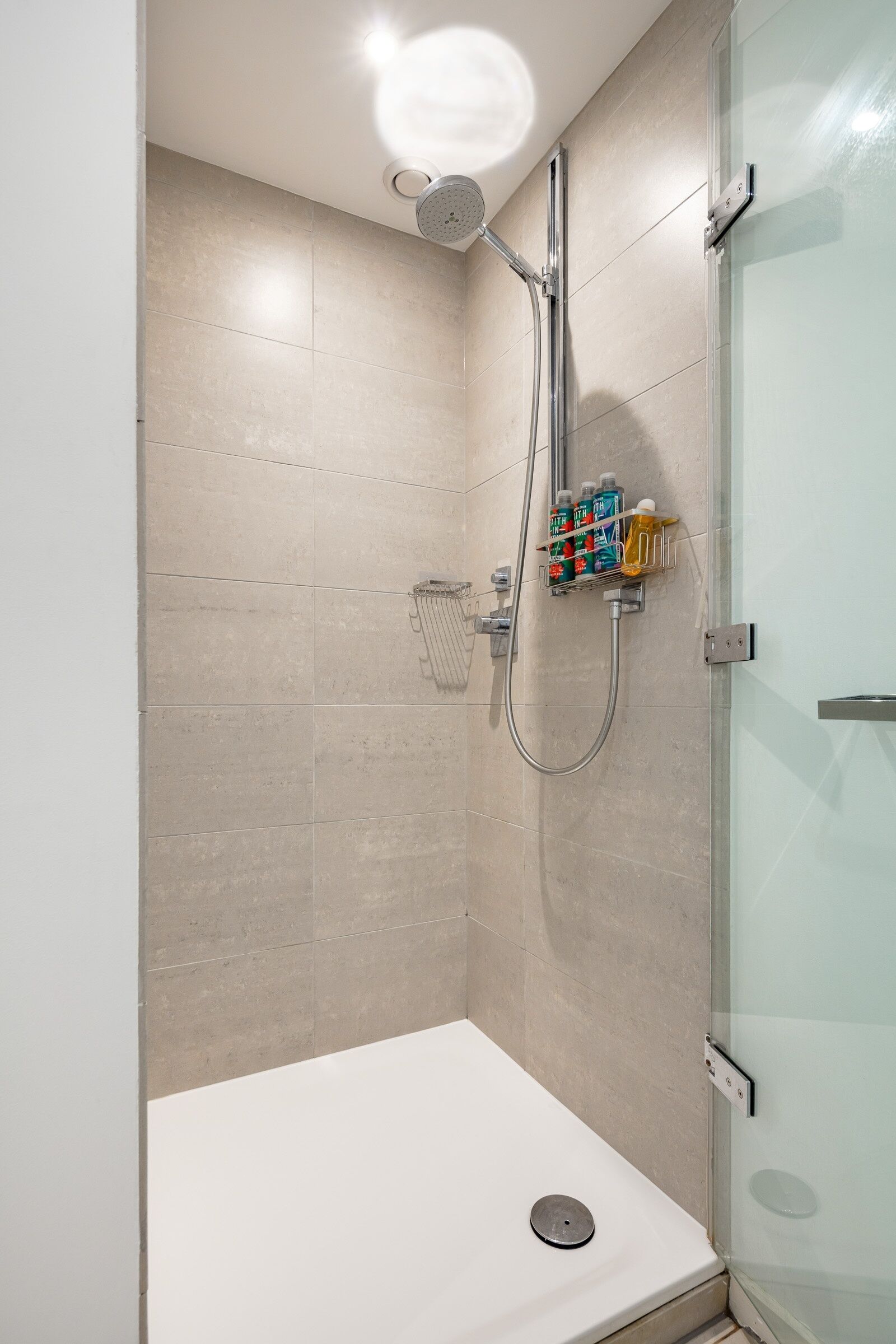Een moderne douche met een glazen deur, beige tegels en een douchekop.