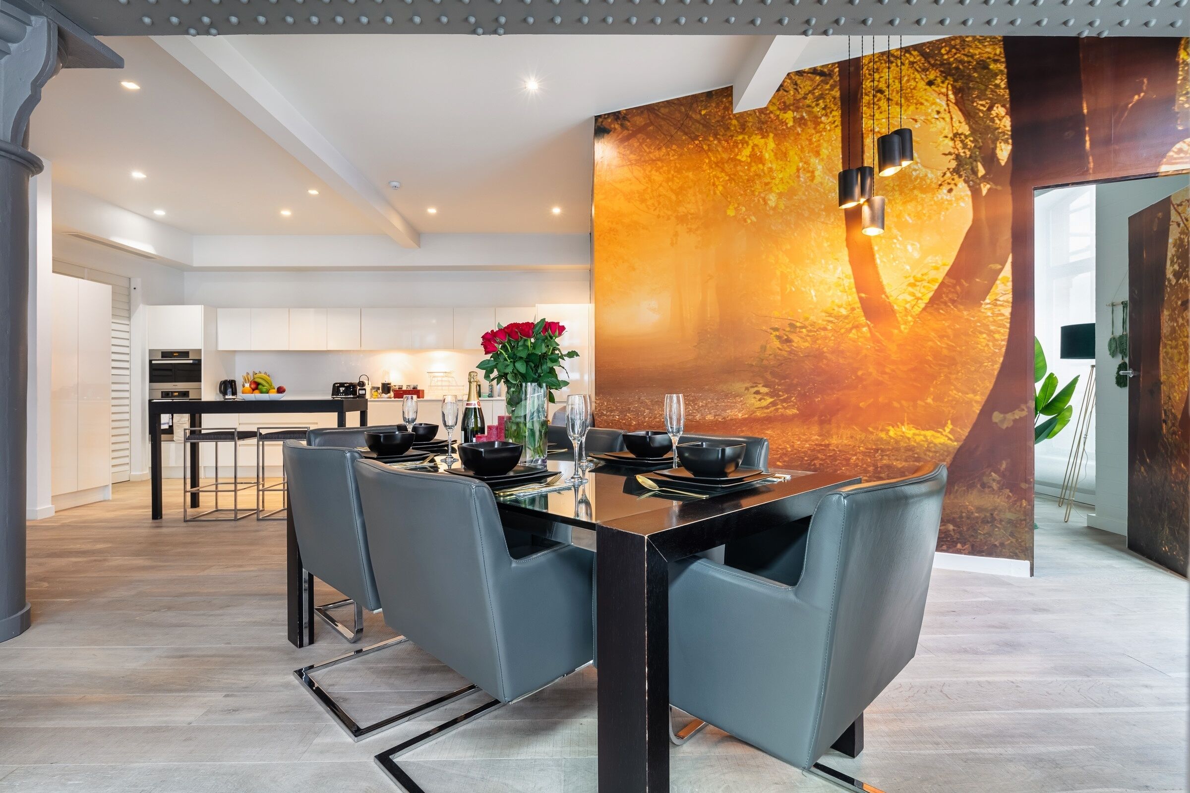 A modern dining area with a glass table, grey chairs, and a decorative wall with a tree mural.