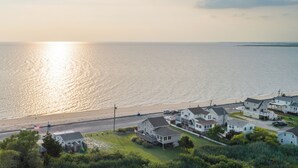 Zona residencial costera con casas a lo largo de una calle frente a la playa, vista despejada del océano y un atardecer de fondo.