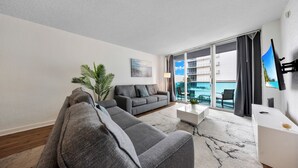 Ein modernes Wohnzimmer mit einem grauen Ecksofa, einem weißen Couchtisch und einem großen Fenster mit Blick auf ein Gebäude und einen Pool.