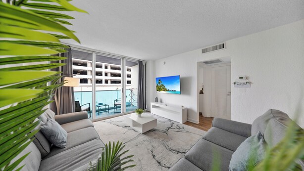 Ein modernes Wohnzimmer mit grauem Sofa, einem weißen Couchtisch und einem an der Wand befestigten Fernseher. Ein Balkon bietet Blick auf ein Gebäude.