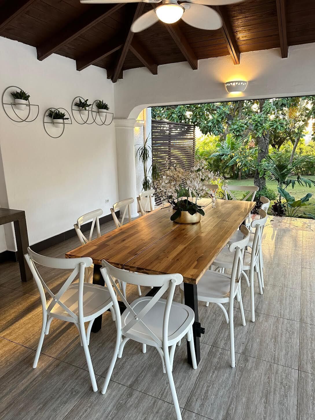 Un coin repas avec une table en bois et des chaises blanches, un ventilateur de plafond et une vue sur un jardin.