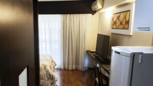 Ein gemütliches Hotelzimmer mit Holzboden, einem Bett mit weißer Bettdecke, einem Schreibtisch mit Computer, einem Fernseher und einem Kühlschrank.
