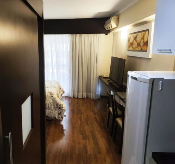 Une chambre d’hôtel compacte avec un plancher en bois, un lit recouvert d’une couette blanche, un bureau avec un ordinateur, une télévision et un réfrigérateur.