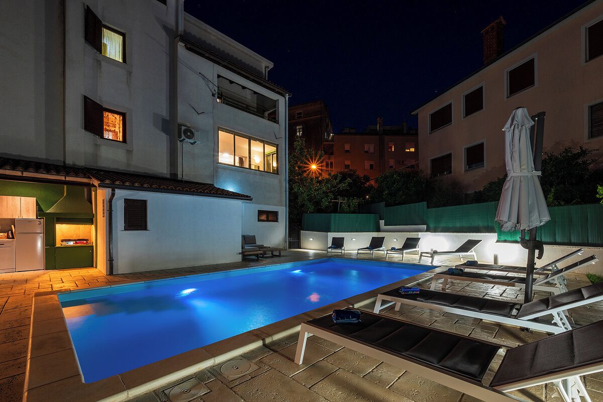 A swimming pool with lounge chairs and umbrellas, surrounded by buildings at night.