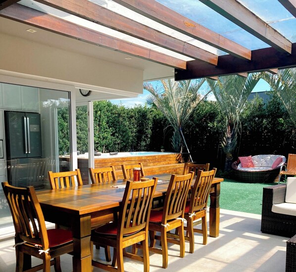 An outdoor dining area with a wooden table and chairs, a glass-enclosed ceiling, and a view of a backyard with a hot tub and greenery. - Rosa Internacional - Surfland (Garopaba)