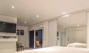 A bedroom with a bed, a TV mounted on the wall, a chair, and a sliding door leading to a balcony. - Rosa Internacional - Surfland (Garopaba)