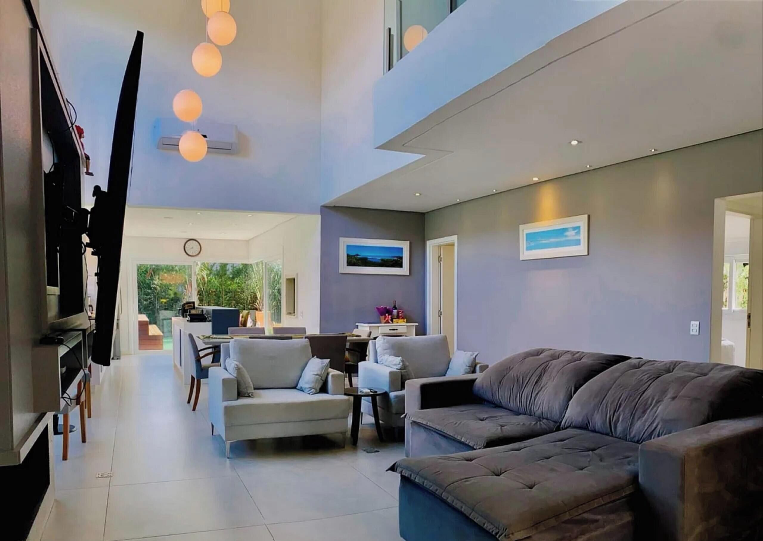 Uma sala de estar moderna com um sofá, poltronas e uma televisão. Ao fundo, há uma área de jantar com uma mesa e cadeiras.