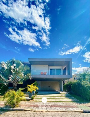 A modern two-story house with a balcony, a paved driveway, and surrounding greenery. - Rosa Internacional - Surfland (Garopaba)