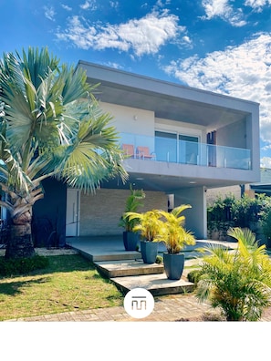 A modern two-story building with a balcony, surrounded by potted plants and a palm tree. - Rosa Internacional - Surfland (Garopaba)