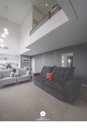 A modern living room with a grey sofa, a black sofa with an orange pillow, and a dining area in the background. - Rosa Internacional - Surfland (Garopaba)