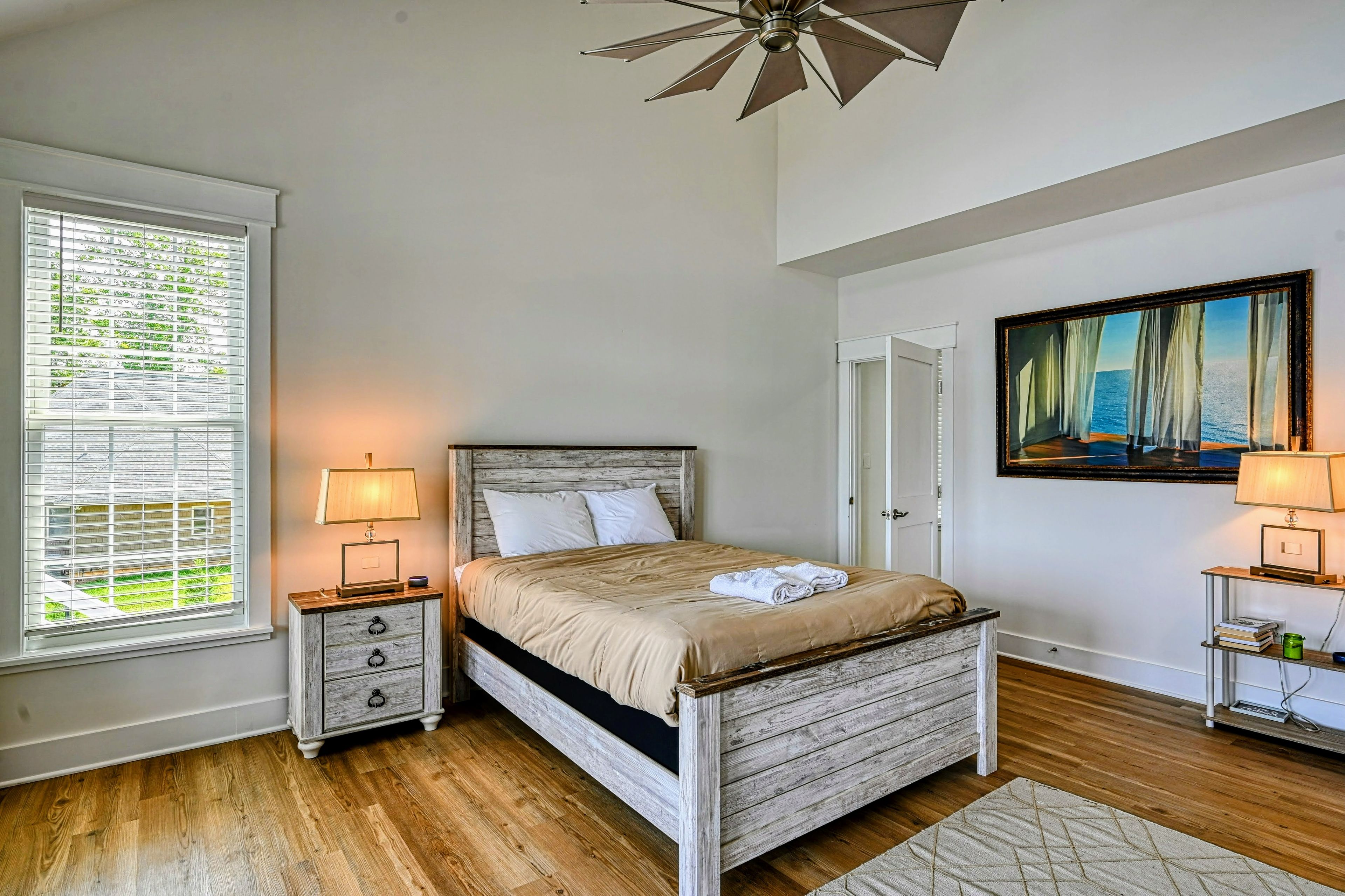 A bedroom with a bed, bedside tables, a ceiling fan, a window with blinds, and a painting on the wall.