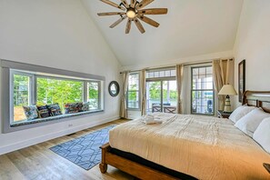 A bedroom with a large bed, a ceiling fan, a window seat with pillows, and a view of greenery outside.