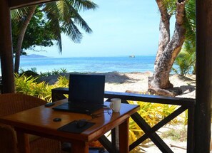 Une table en bois avec un ordinateur portable et une tasse, donnant sur une plage bordée de palmiers et sur l’océan.