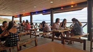 An outdoor dining area with wooden tables and benches, overlooking a body of water and hills. - Ratu Kini's Dive & Stay (Mana Island)
