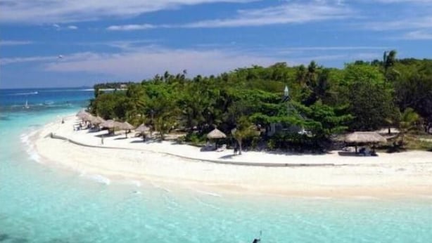A tropical beach with clear turquoise water, a sandy shore, and a small island with lush greenery and thatched-roof structures. - Ratu Kini's Dive & Stay (Mana Island)