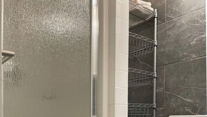 A bathroom with a glass shower door, a white tiled wall, a black trash bin, and a metal shelf unit. - Great Mid-Town Toronto Bachelor Suite Convenient to Downtown and Airport (Toronto)