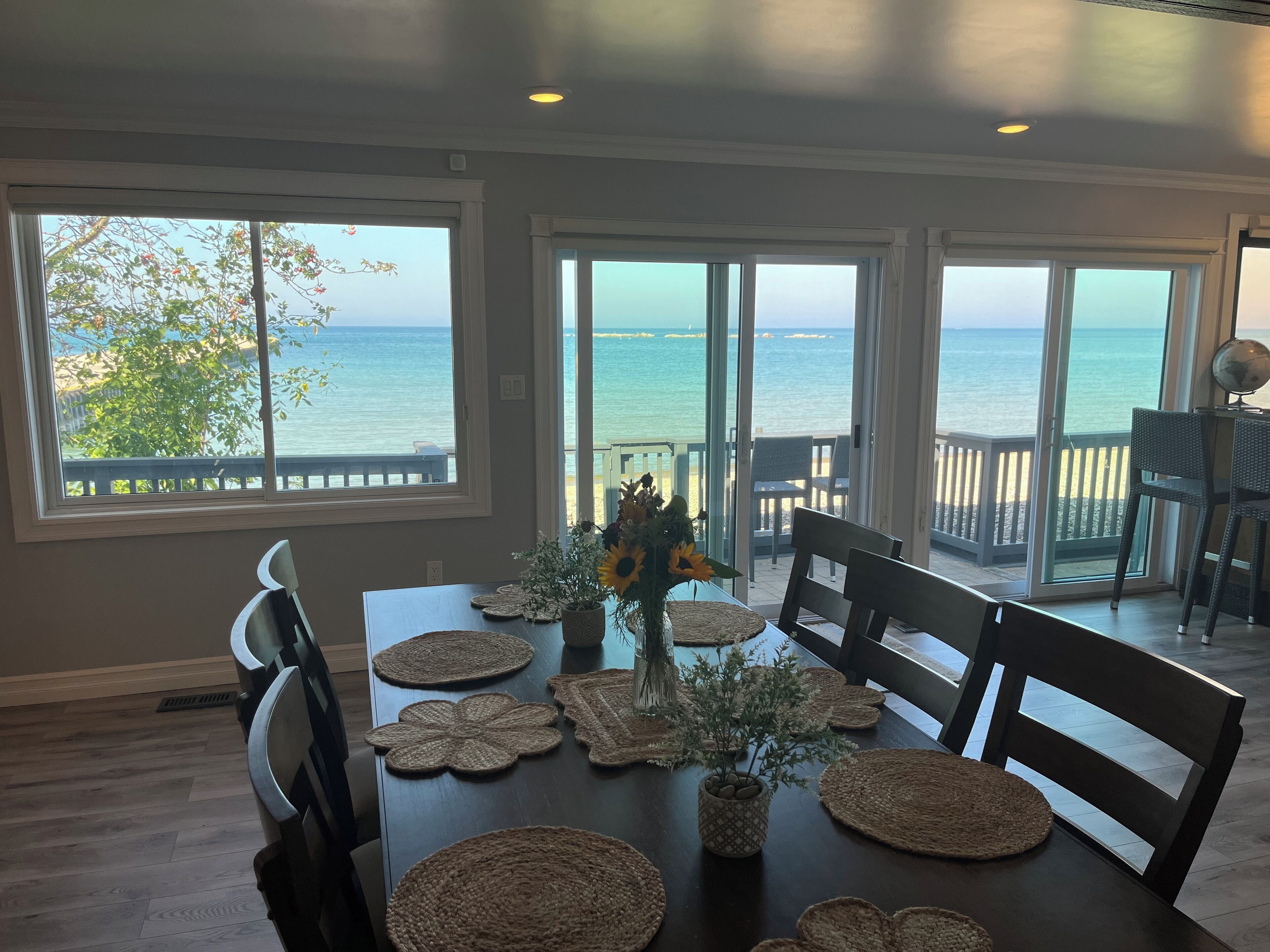 A dining area with a view of the ocean, a table set with placemats and a vase of flowers, and a chair with a globe on it.