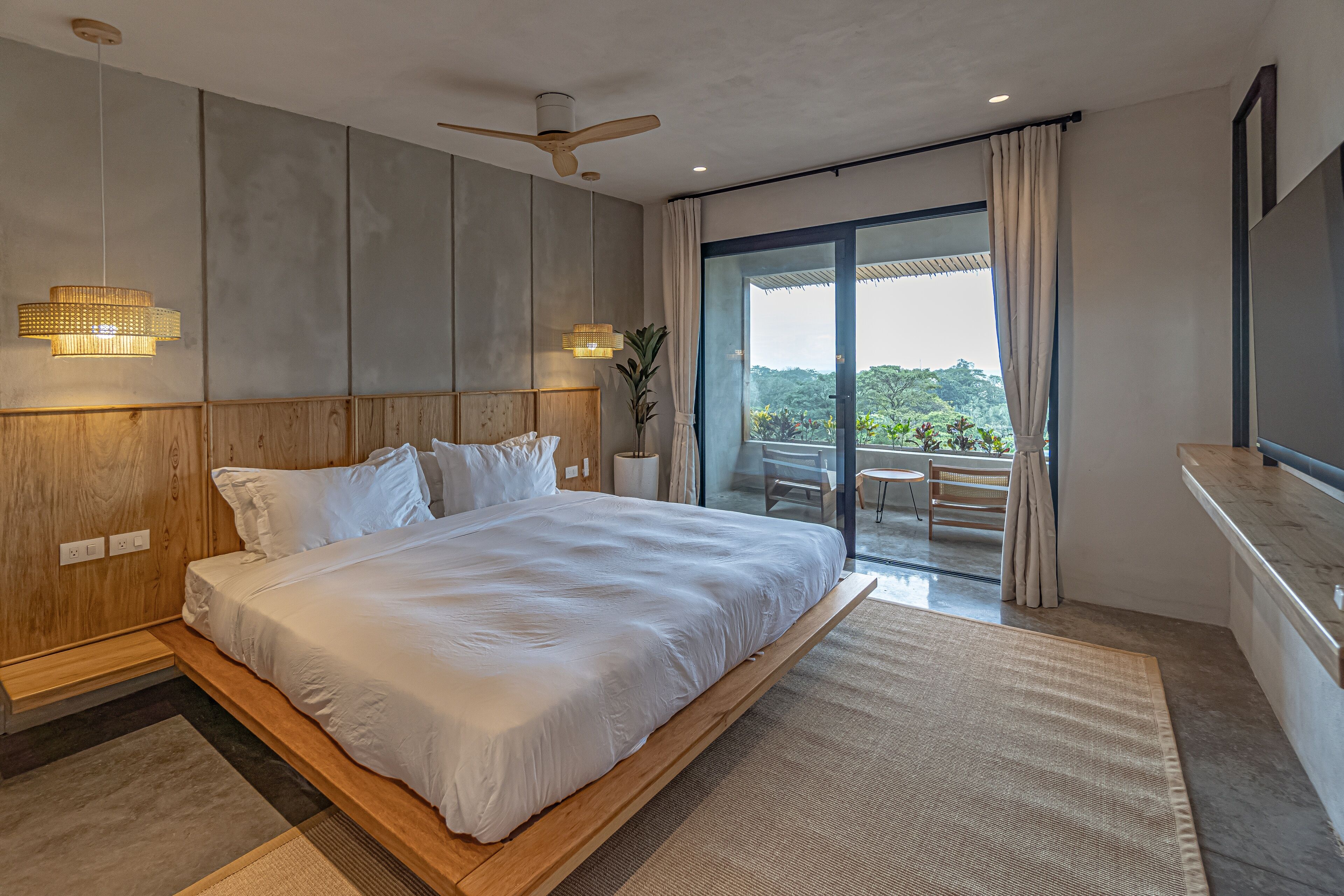 Un dormitorio moderno con una cama grande, cabecera de madera, ventilador de techo y vistas al exterior a través de un ventanal corredero.