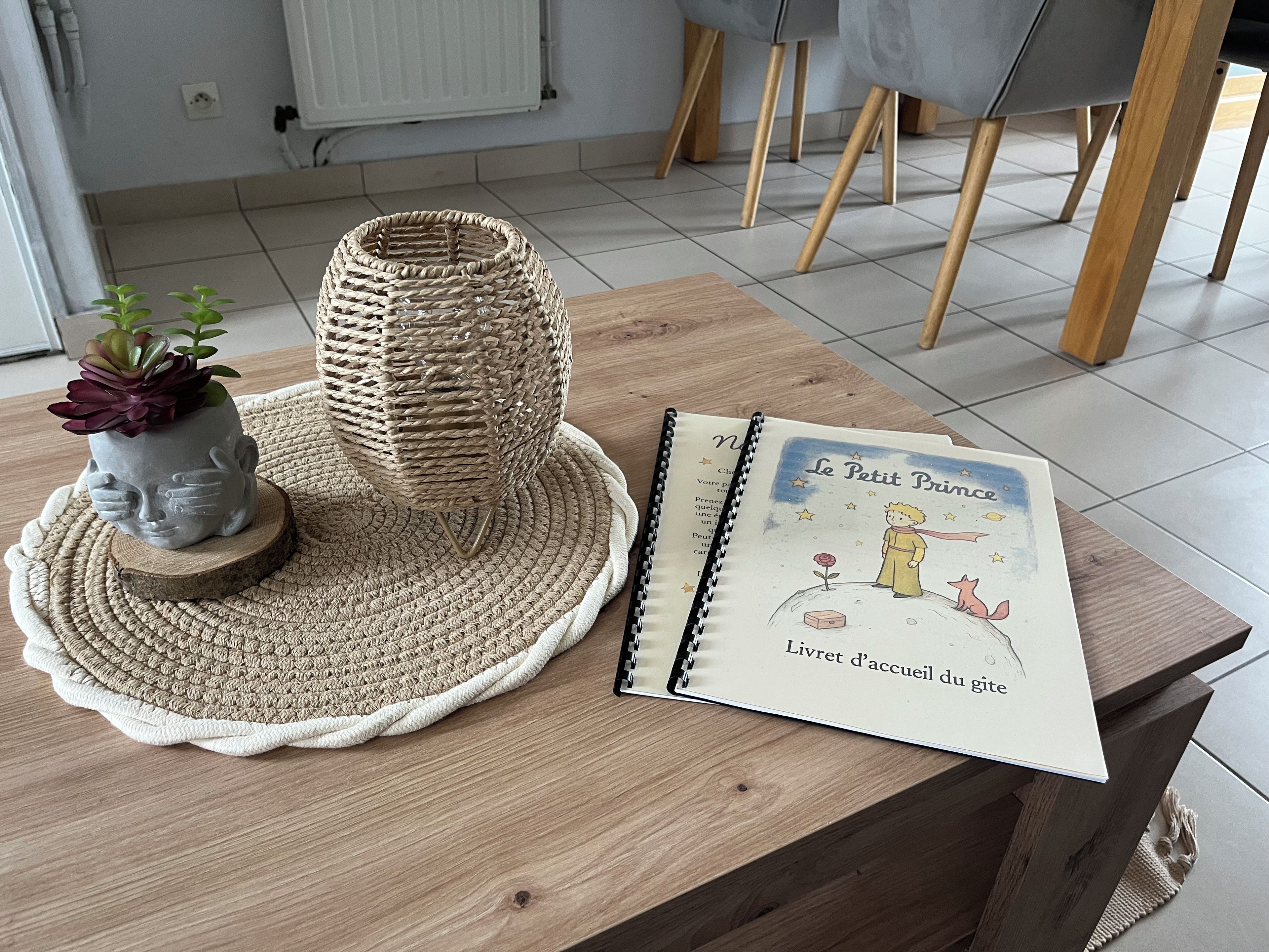 A wooden table with a woven basket, a ceramic planter with a succulent, and a book titled 'Le Petit Prince' on a woven placemat.