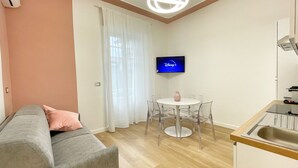 Ein kleiner, moderner Wohnraum mit einem grauen Sofa, einem runden Tisch mit Stühlen, einem an der Wand montierten Flachbildfernseher und einer Küchenzeile mit Spüle und Arbeitsplatte.