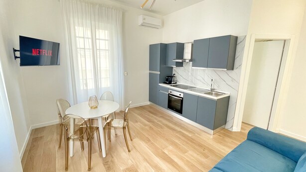 Ein kompakter Wohnbereich mit Küchenzeile, einem Essbereich mit Tisch und Stühlen, einem blauen Sofa und einem an der Wand befestigten Fernseher.