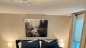 Una cama grande con ropa oscura, dos lámparas de noche, un cuadro en la pared y una alfombra estampada.