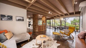 Een ruime woonkamer met een houten plafond, een eethoek met gele stoelen, en een keuken die door een open deur zichtbaar is.
