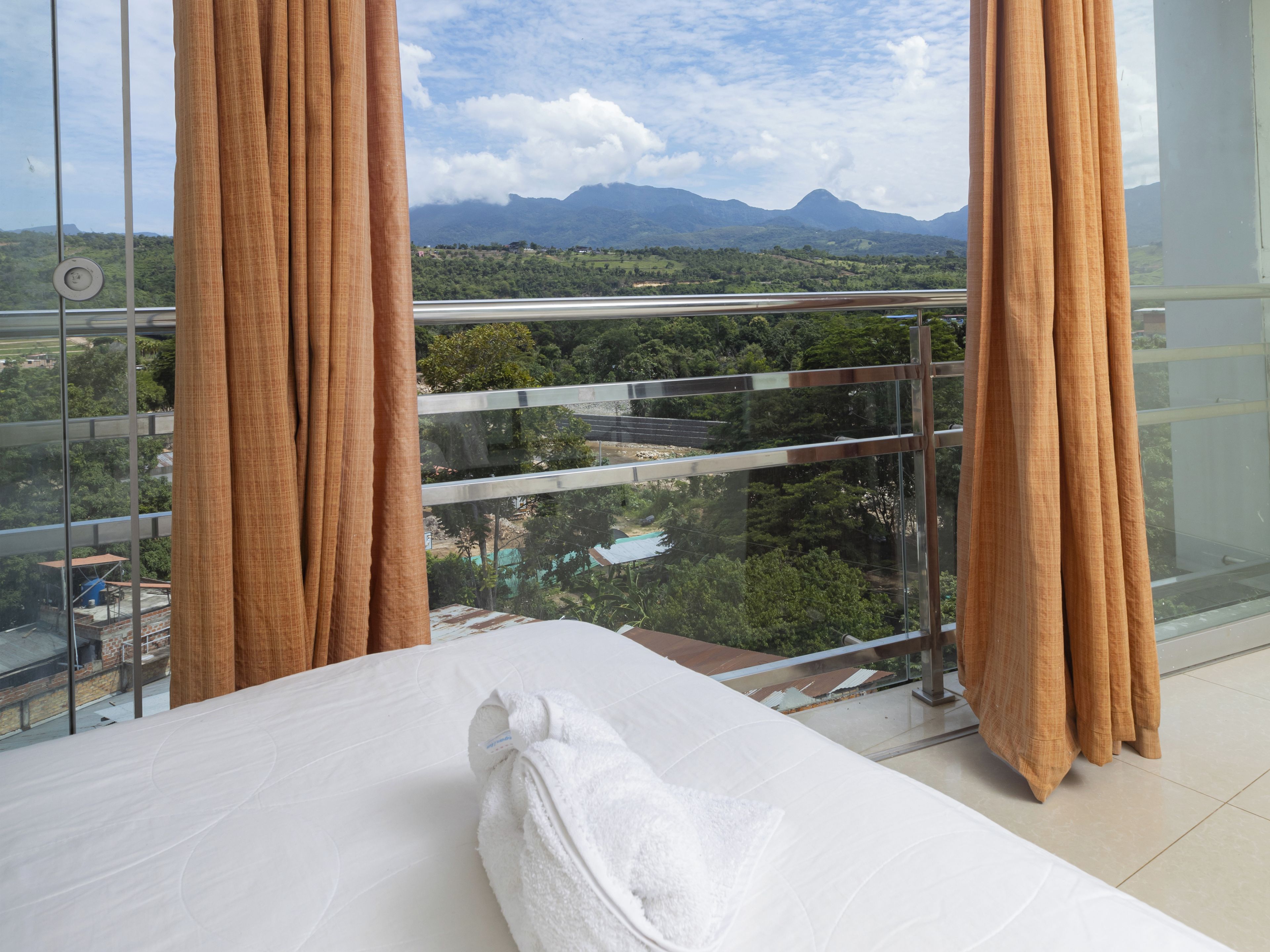 A hotel room with a bed, curtains, and a view of a mountainous landscape through a glass balcony.