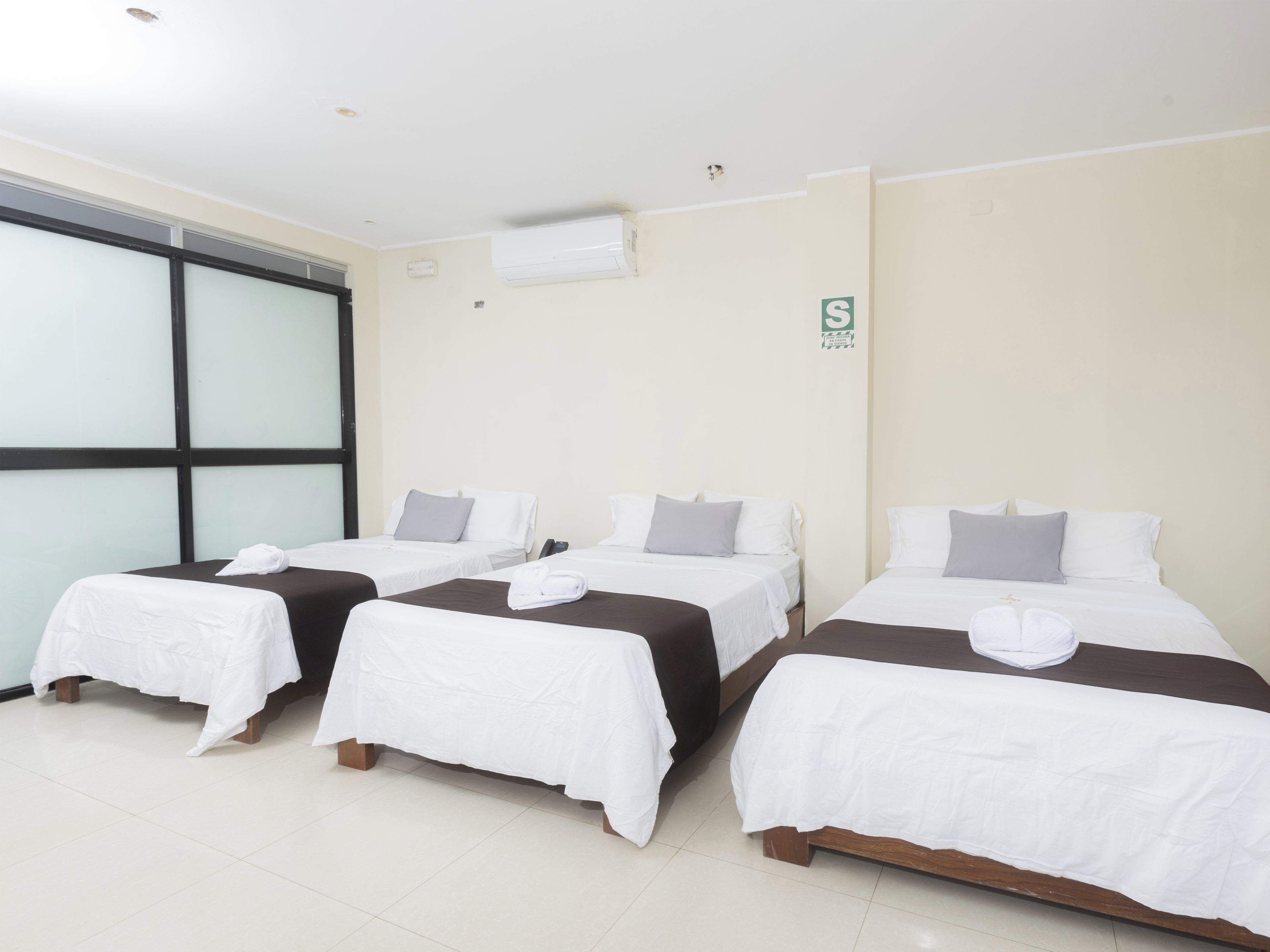 A hotel room with three single beds, each with a white bedspread and a grey pillow. There is a window with blinds, an air conditioning unit on the wall, and a sign with the letter 'S'.