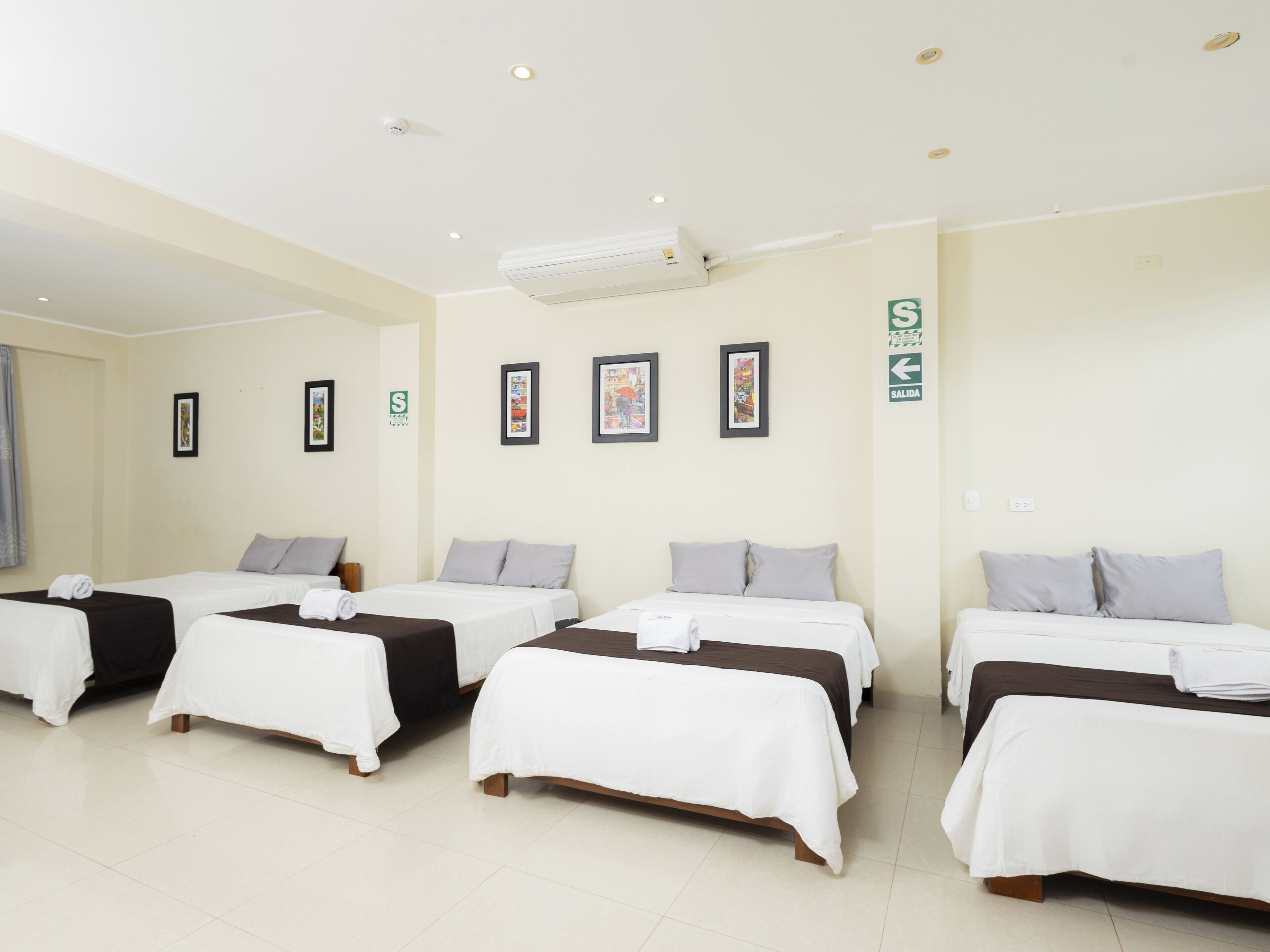 Habitación de hotel con cuatro camas dispuestas en fila, cada una con cubrecama blanco y almohadas grises. Hay cuadros enmarcados en la pared, un aire acondicionado en el techo y una señal de salida verde.