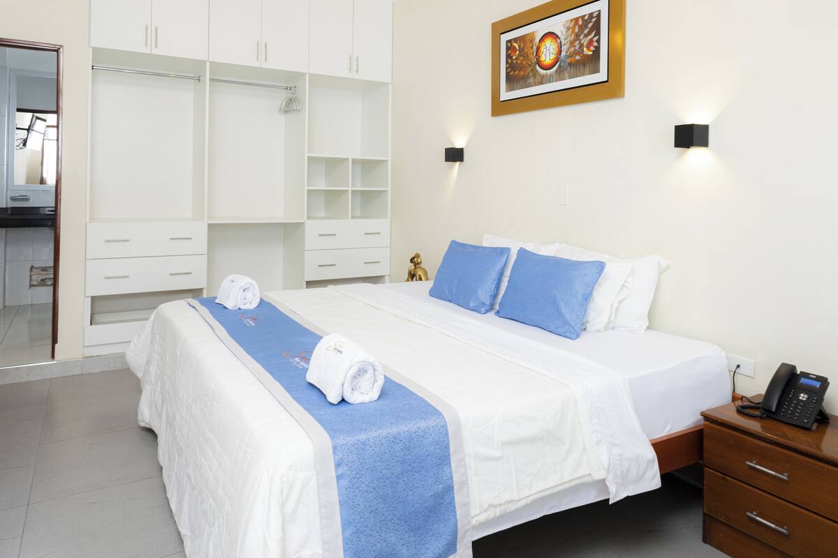 Habitación de hotel con una cama grande, una mesita de noche de madera, un teléfono y un armario con varios compartimentos.
